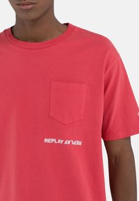 Punainen puuvillapaita pyöreällä pääntiellä, lyhyillä hihoilla ja rintataskulla. Ominaisuuksina valkoinen "REPLAY" -logoteksti alaosassa edessä.