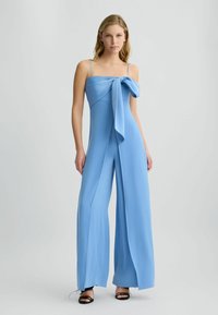 Blaues Jumpsuit mit einer gebundenen Schleifendetail am Ausschnitt, Spaghettiträgern, weitem Bein und weicher Textur. Schwarze Sandalen runden den Look ab.