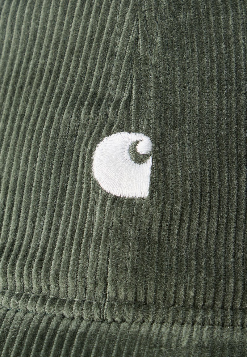 Tessuto in velluto a coste verde con logo curvo ricamato in bianco su un motivo a coste verticali testurizzate.