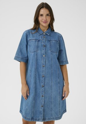 Donna che indossa un vestito di jeans blu di lunghezza media con maniche corte e chiusura frontale con bottoni, in piedi su uno sfondo bianco uniforme.