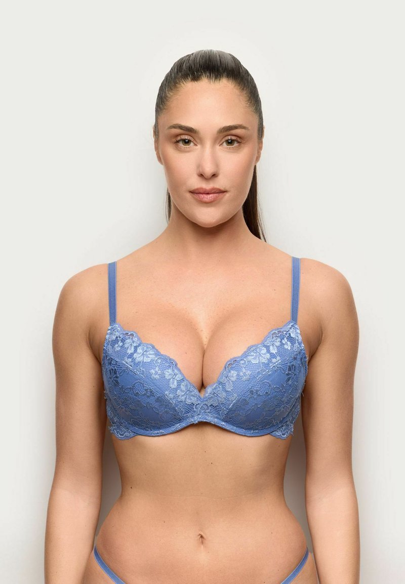 Vrouw met lang donker haar in een blauw kanten beha, staand tegen een effen lichte achtergrond, voorwaarts gericht met een neutrale uitdrukking.