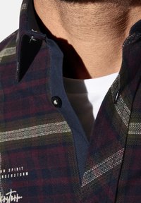 Knopfhemd mit dunklem Karomuster in Burgunderrot, Grün und Marineblau; mit Kragen und weißem Unterhemd, das darunter sichtbar ist.