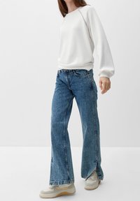 White sweatshirt with ribbed cuffs, blue high-waisted wide-leg jeans with raw hems, and light-colored sneakers with layered details.