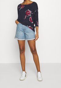 Mörkblå långärmad skjorta med röd blommönster, tillsammans med ljusa denimshorts. Modellen bär vita sneakers och står mot en enkel bakgrund.