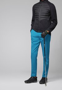 Černá prošívaná bunda na zip, světle modré kalhoty a černé golfové boty. Drží golfovou hůl a nosí černé rukavice se zelenými akcenty.