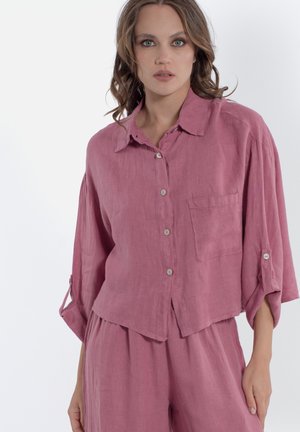 Donna che indossa una camicia color malva a manica lunga con taschino sul petto e pantaloni a vita alta e gamba larga abbinati, in piedi su sfondo bianco.