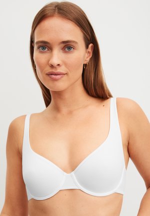 Donna con capelli castano chiaro lisci, che indossa un reggiseno bianco con ferretto e piccoli orecchini a cerchio dorati, su sfondo chiaro.
