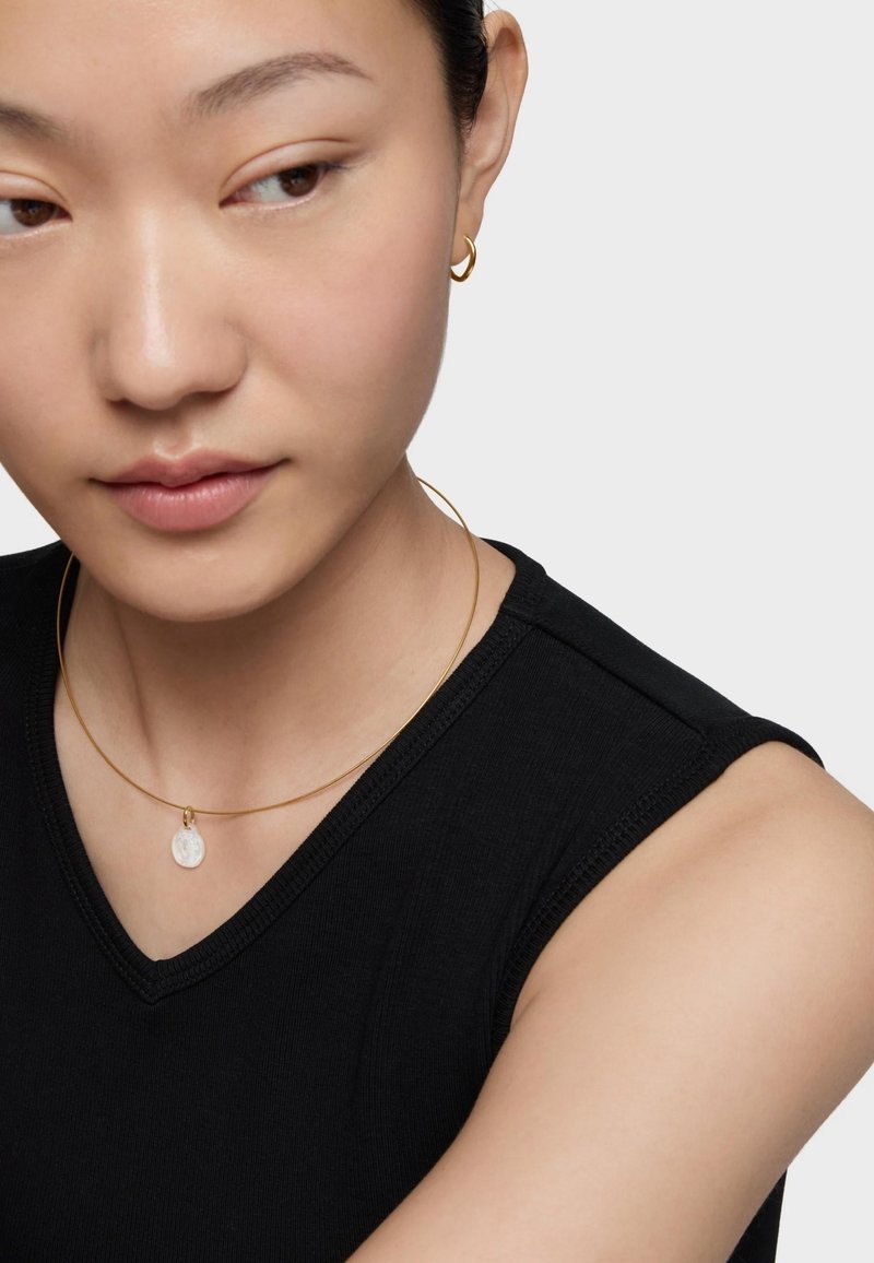 Joyería en tono dorado que incluye un collar con cadena delgada y un colgante redondo, junto con pendientes de aro a juego, todo ello sobre una blusa de tirantes negra.