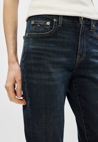 Granatowe dżinsy z klasycznym pięciopocketowym krojem, z widocznym przeszyciem, mosiężnymi detalami i małym logo na przedniej kieszeni.