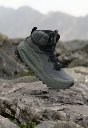 Botte de randonnée grise et noire en l'air au-dessus d'un terrain rocheux avec des montagnes floues en arrière-plan.