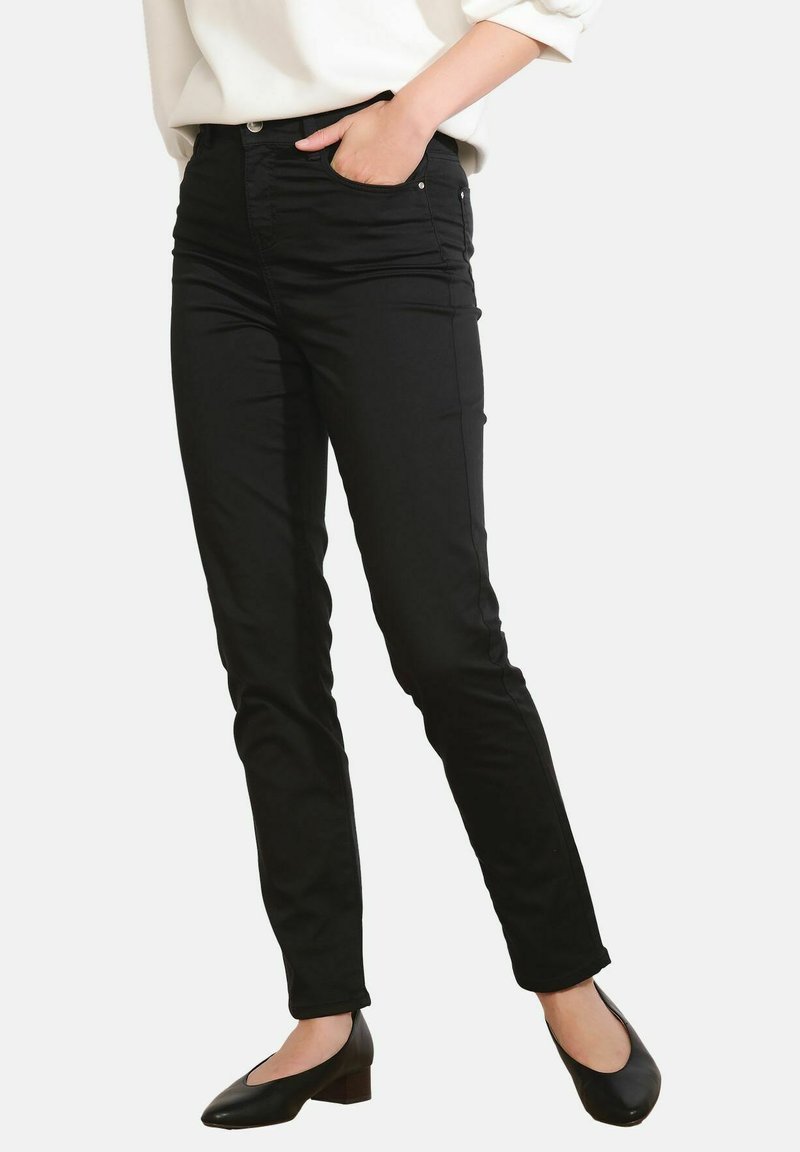 Persona che indossa pantaloni neri skinny con una mano in tasca e scarpe nere con tacco basso, abbinati a una maglietta bianca a maniche lunghe.