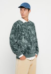 Pūkains zaļš džemperis ar teksturētu rakstu, ribinātām piedurknēm un apakšmalu, virs baltas krekla ar apkakli, apvienots ar zilu džinsa cepuri.