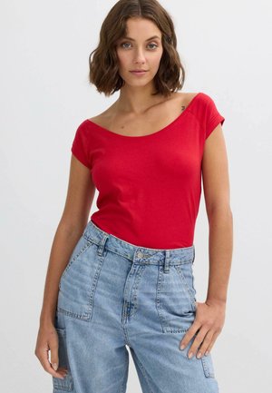 Mujer con cabello castaño ondulado, lleva una blusa roja sin hombros y vaqueros azul claro de talle alto, de pie frente a un fondo blanco liso.