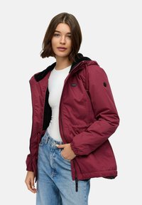 Giacca bordeaux con zip, cappuccio foderato in pile nero, polsini elasticizzati e coulisse in vita, indossata sopra una camicia bianca.