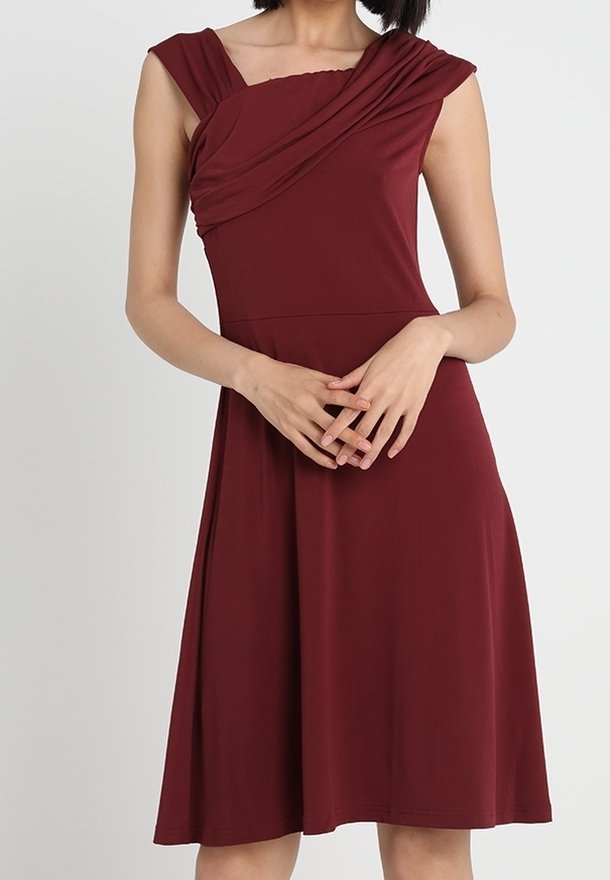 Femme portant une robe sans manches, longueur genoux, rouge foncé avec des détails plissés sur le haut du corsage et les mains croisées à la taille.