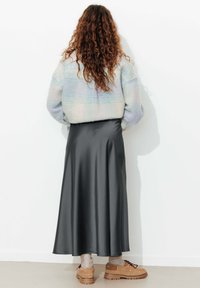 Femme avec de longs cheveux bouclés portant une veste courte pastel, une jupe midi en satin noir, des chaussettes beiges et des chaussures à lacets marron, se tenant contre un mur blanc.