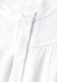 Camicia bianca con tessuto testurizzato e stropicciato, scollo rotondo e quattro bottoni abbinati lungo la patta per la chiusura.