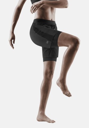 Personne en shorts de sport noirs et crop top levant le genou gauche, en équilibre sur le pied droit devant un fond uni clair.