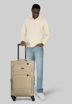 Mann in beigefarbenem Langarmshirt und Jeans, steht mit einer Hand in der Tasche und hält mit der anderen einen beigen Rollkoffer.