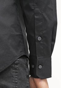 Camisa negra de botonadura con mangas, textura suave, detalle de puño con dos botones y un dobladillo ligeramente curvado, combinada con jeans de mezclilla oscura.