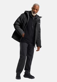 Giacca nera impermeabile con cappuccio, chiusura a zip e tasche laterali; pile nero sotto, abbinato a pantaloni neri e sneakers.