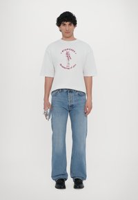 White oversized t-shirt featuring red graphic print, paired with light blue jeans. Model holds a small silver object in one hand.