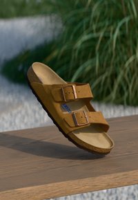 Sandal com palmilha de camurça castanha, com duas tiras ajustáveis, fivelas metálicas e uma base de cortiça. Sola texturizada preta. Design plano.