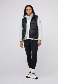 Schwarze Steppweste über einem hellen Mint-Fleece-Oberteil, kombiniert mit schwarzen Leggings und weißen Schuhen mit orangefarbenen Akzenten. Einfacher Hintergrund.