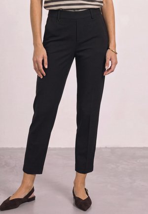 Femme portant un pantalon court noir droit, un haut rayé, des ballerines marron à bout pointu avec bride à l'arrière, et un bracelet en or, debout sur un sol gris.