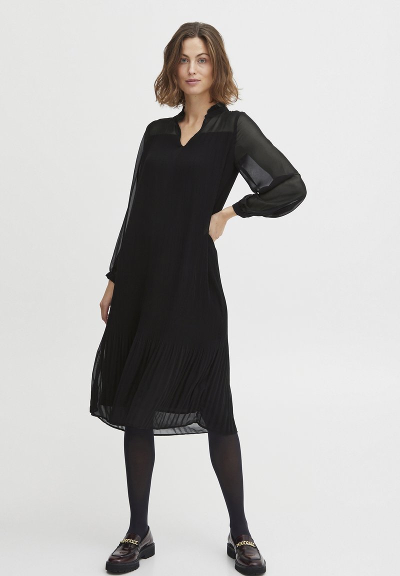 Mustat pleated-mekko, jossa on läpinäkyvät pitkät hihat, V-pääntie ja väreilevä siluetti, yhdistettynä tummaan sukkahousuun ja loferi-kenkiin, joissa on ketjutehosteita.