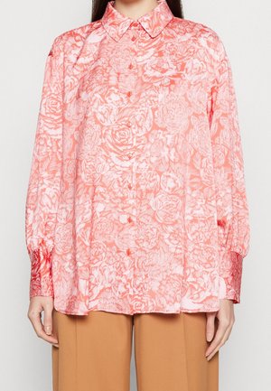 Kvinde iført en koralrosa blomster-knap-op skjorte med lange ærmer, kombineret med lysebrune bukser, stående mod en ensfarvet baggrund.