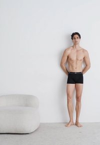 Boxers noirs avec une texture lisse et une taille élastique, portés par une silhouette masculine debout sur un fond clair.