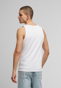 White cotton tank top with a round neck and wide armholes, viewed from the back. Paired with light blue jeans. Minimal design.