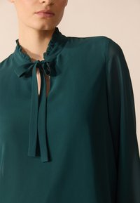 Teal blouse with a ruffled collar and a bow tie detail at the neckline, made from a smooth, lightweight fabric. Long sleeves are slightly sheer.