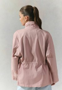 Veste en coton rose clair avec un col montant, taille élastique et manches larges, affichant une couleur unie sans motifs ni accents visibles.