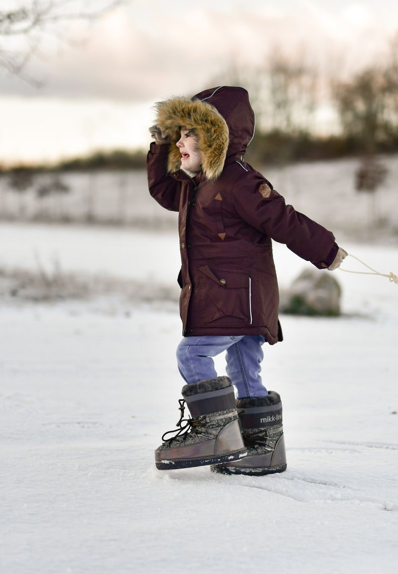 Ruda parka su kailio apdaila ant gobtuvo, violetinės kelnės ir pilki žieminiai batai. Lauko snieguotas vaizdas su vaiku, kylančiu ranką.