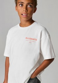 Bílý krátký rukáv tričko s nápisem "ALLSAINTS" a dalším textem v korálové barvě na hrudi, volný střih, vyrobeno z bavlněné tkaniny.
