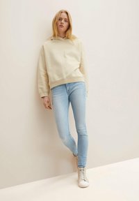 Beige huva sweatshirt, ljusblå slim fit jeans och grå höga sneakers; mjuk textur, avslappnad design och åtsittande silhuett.