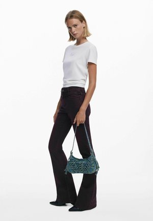 Fehér pólót és sötét bővülő farmert viselő nő, kék textúrájú táskával a kezében, egy sima fehér háttér előtt áll.