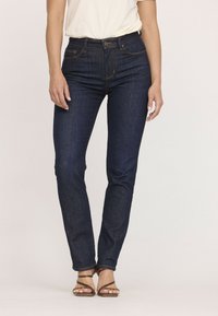 Jean en denim bleu foncé avec une coupe slim, taille haute et couture orange contrastante. D'autres détails incluent des poches et une fermeture à bouton.