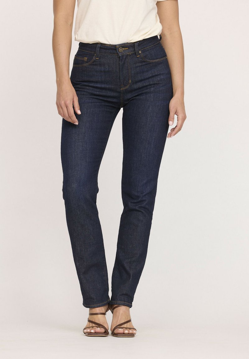 Jean en denim bleu foncé avec une coupe slim, taille haute et couture orange contrastante. D'autres détails incluent des poches et une fermeture à bouton.