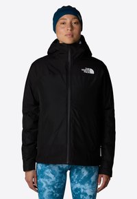 Czarna ocieplana kurtka z kapturem zapinana na zamek, z białym logo na lewej piersi. Nosiiona na niebieskie legginsy w wzory.