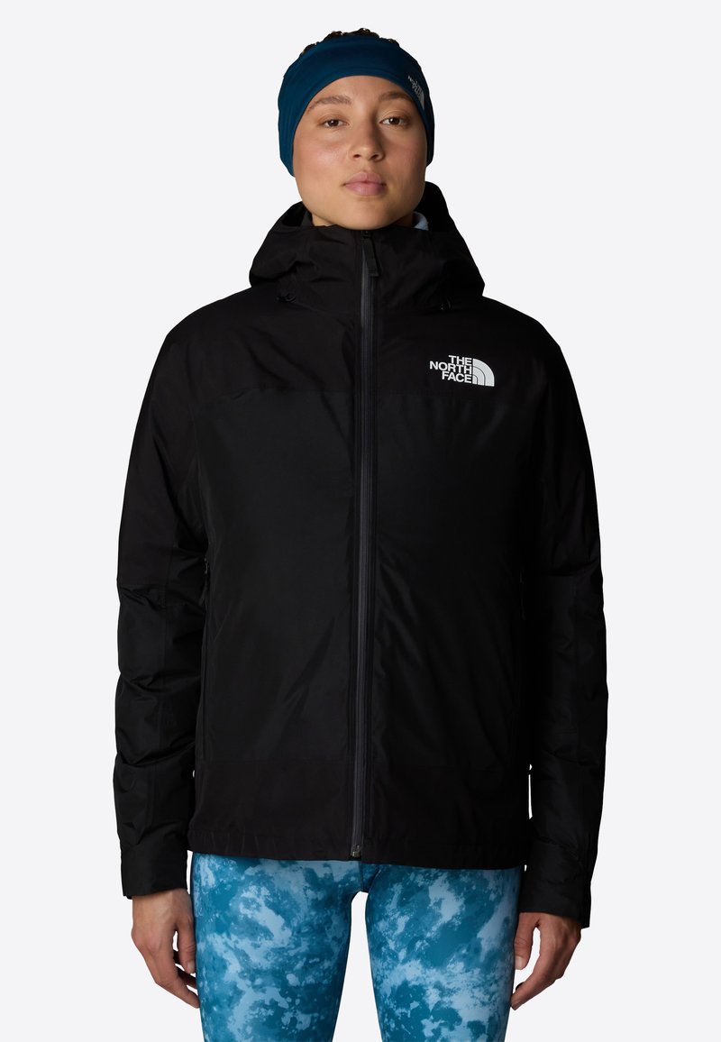 Czarna ocieplana kurtka z kapturem zapinana na zamek, z białym logo na lewej piersi. Nosiiona na niebieskie legginsy w wzory.