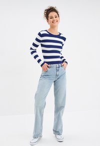 Gestreiftes, dunkelblaues und weißes, geripptes Langarmoberteil, kombiniert mit hellblauen, weiten Jeans und weißen Turnschuhen. Das Model posiert mit den Händen in den Hüften.