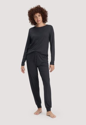 Frau mit lockigem Haar, die ein schwarzes langärmliges Oberteil und schwarze, mit Knöpfen versehene Freizeithosen trägt, steht barfuß vor einem hellen Hintergrund.