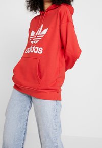 Person med röd Adidas-hoodie med vitt logotyp, kombinerad med ljusblå jeans, stående mot en enfärgad vit bakgrund.