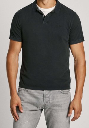 Man die een donkerblauw poloshirt draagt over een wit ondershirt en lichtgrijze spijkerbroek, staand tegen een effen achtergrond.