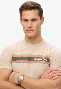 Camiseta de algodón beige con franjas verdes, naranjas y moradas, con el logo de "SuperDry". El modelo lleva una reloj de tono dorado con una correa de cuero marrón.