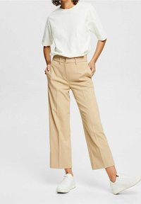 Personne portant un pantalon beige taille haute à jambes larges, un haut blanc à manches courtes et des baskets blanches à plateforme, debout les mains dans les poches.