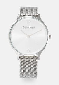 Strieborné hodinky na sieťovom remienku s minimalistickým okrúhlym ciferníkom. Biely ciferník obsahuje strieborné hodinové značky a logo "Calvin Klein" na pozícii 12.